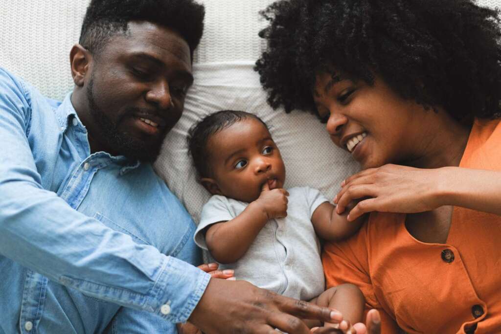 Father and mother lay on bed with baby laying between them; Benefits of paternity leave
