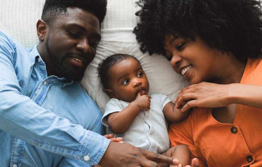 Father and mother lay on bed with baby laying between them.