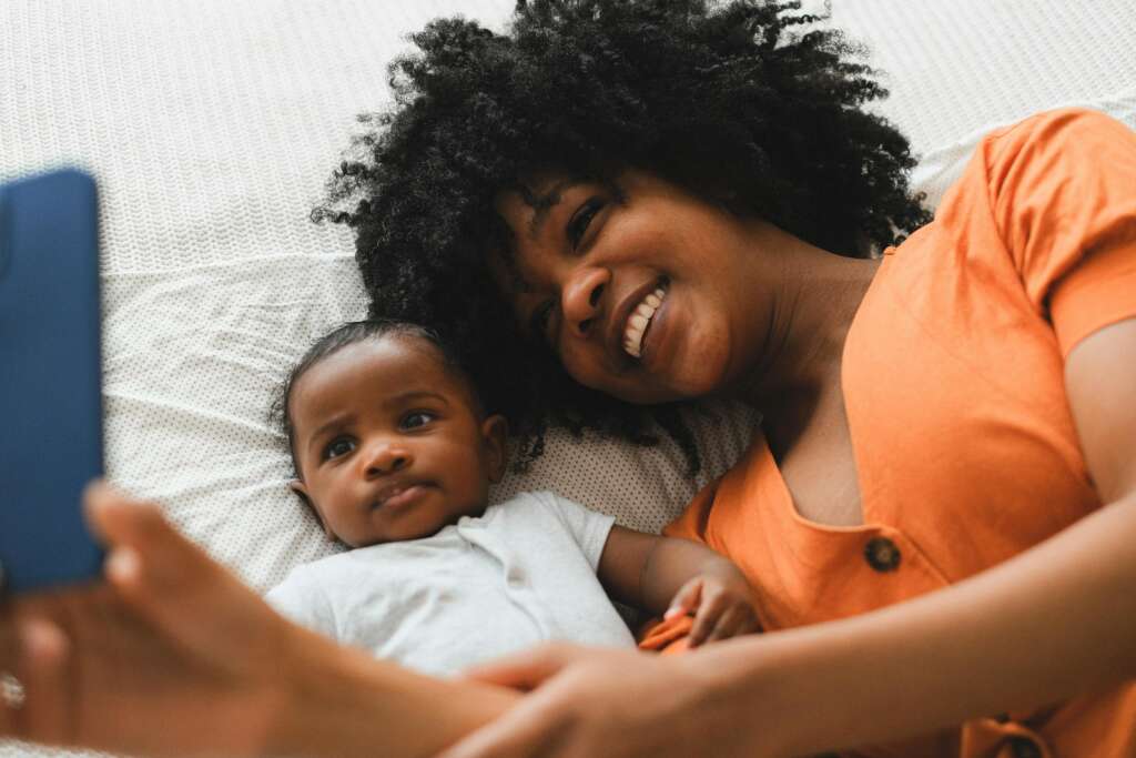 Happy mother and baby lying on bed taking a selfie, smiling joyfully; sharenting and your child's digital footprint